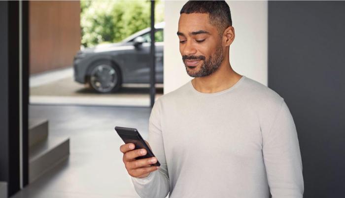 Ein Mann blickt auf sein Smartphone. Im Hintergrund durch eine Glasscheibe zu erkennen: ein Audi Modell, das vor seinem Zuhause parkt.