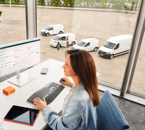 Das Bild zeigt einen Büroarbeitsplatz, an dem eine Fuhrparkmanagerin vor einem Computer sitzt und auf den Bildschirm schaut. Im Hintergrund sind der neue Transporter, ein ID. Buzz Cargo, ein Caddy Cargo und ein Crafter zu sehen.