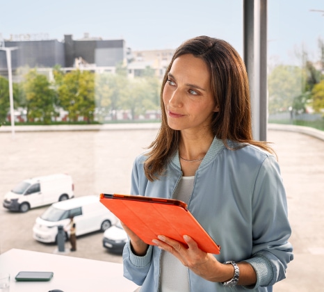 Das Bild zeigt einen Büroarbeitsplatz, an dem eine Fuhrparkmanagerin mit einem orangenen Tablet arbeitet. Sie schaut nach vorne. Im Hintergrund sind vier verschiedene Volkswagen Nutzfahrzeuge zu sehen: der neue Transporter, ein ID. Buzz Cargo, ein Caddy Cargo und ein Crafter.
