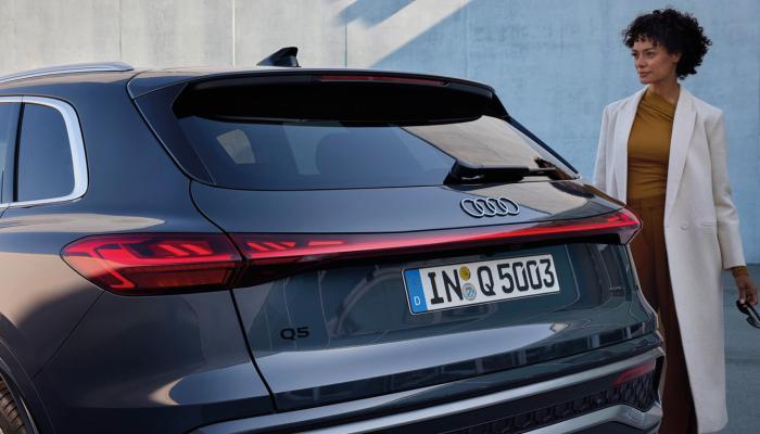 Eine Fahrerin des neuen Audi Q5 vor ihrem Fahrzeug. Zu sehen ist das Fahrzeugheck mit den OLED-Heckleuchten.