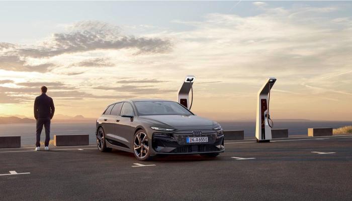 Ein Audi A6 Avant e-tron an einer Schnellladestation. Sein Fahrer steht neben dem Fahrzeug und genießt den Ausblick auf eine Bergkette im Abendrot.