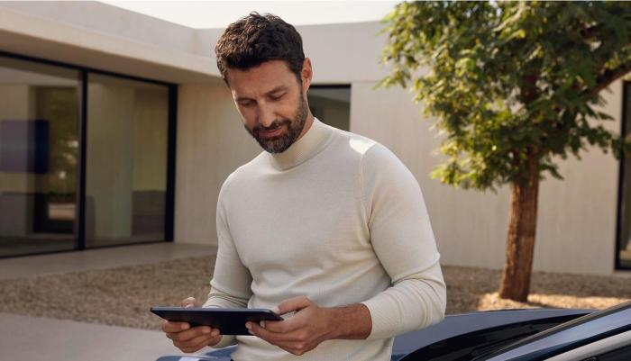 Ein Audi Fahrer lehnt entspannt an seinem Fahrzeug und bedient sein Tablet. Die Szenerie ist in mildes Sommerlicht getaucht. Im Hintergrund ein modernes, flaches Wohngebäude in Terracotta-Tönen.