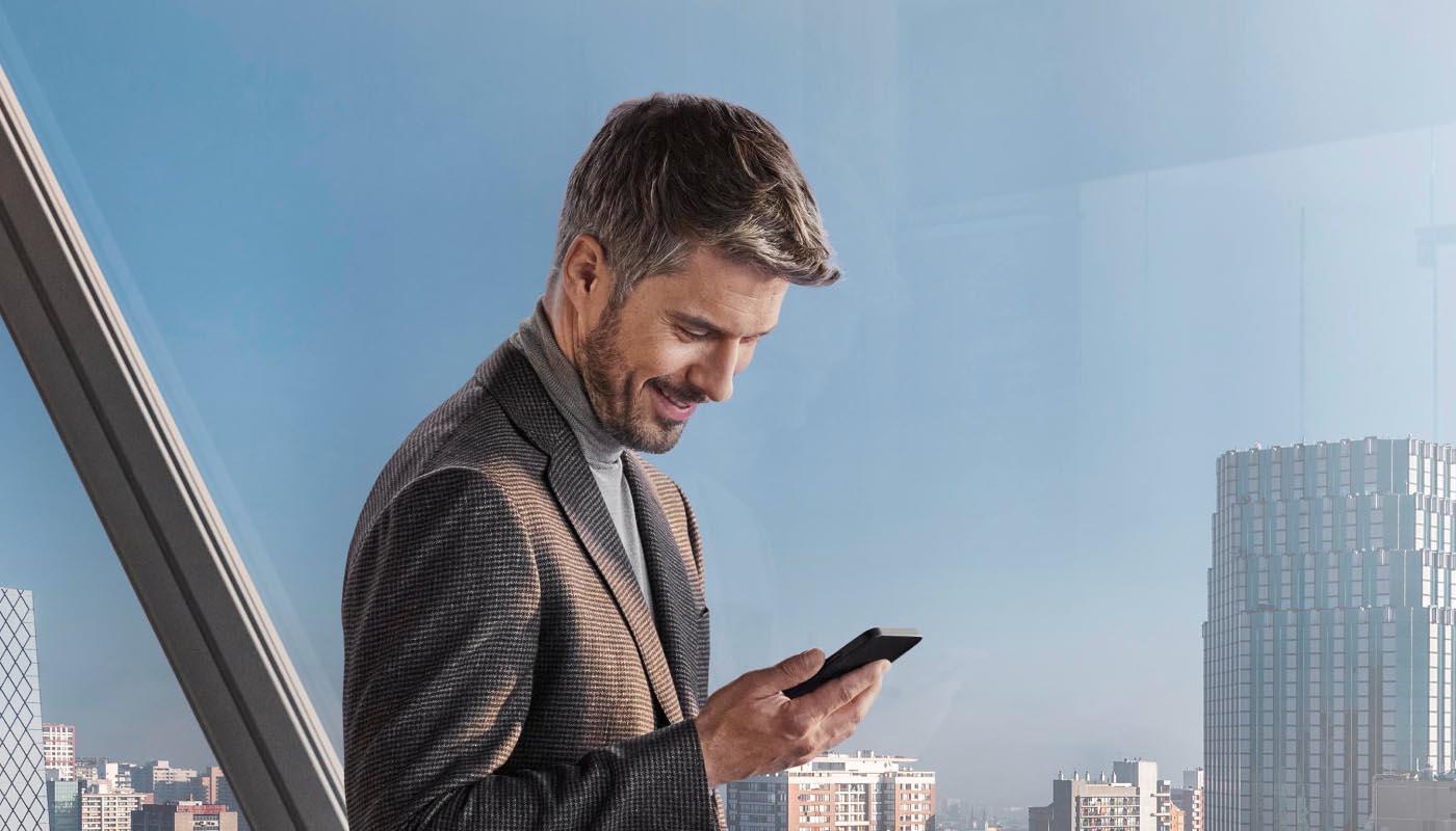 Ein Mann in Business-Kleidung blickt auf sein Smartphone. Im Hintergrund: eine urbane Skyline.