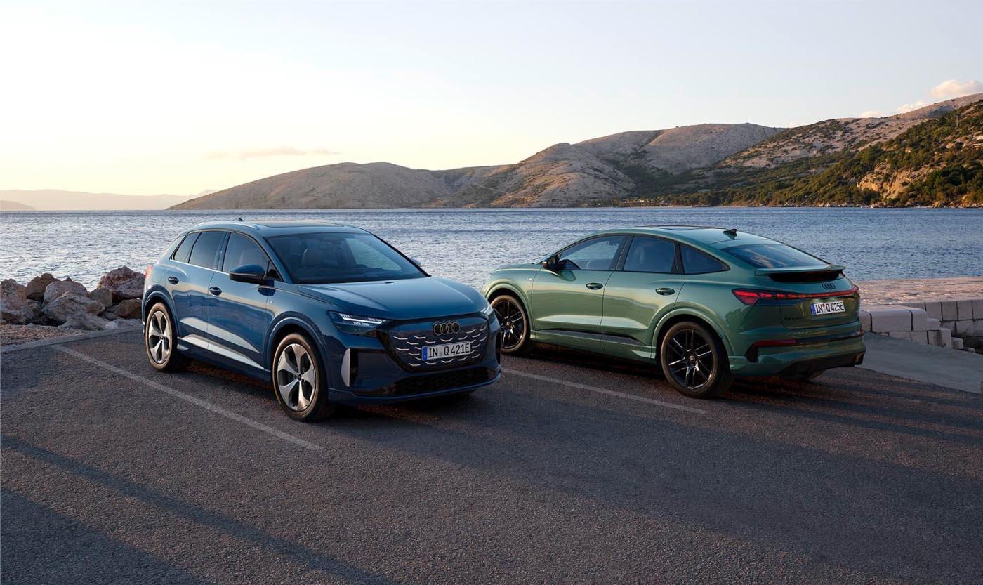 Sanfte Abendstimmung: Ein Audi Q4 e-tron SUV in Plasmablau Metallic und ein Audi Q4 e-tron Sportback in Salbeigrün Metallic parken am Ufer einer Meeresbucht.