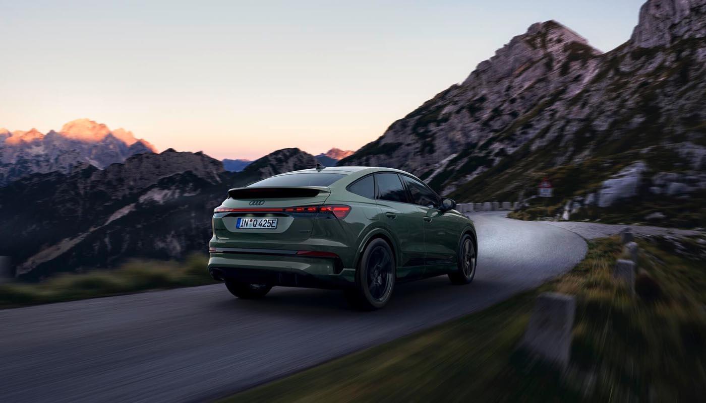 Ein Audi Q4 e-tron Sportback in Salbeigrün Metallic fährt bei Sonnenuntergang durch eine Berglandschaft.