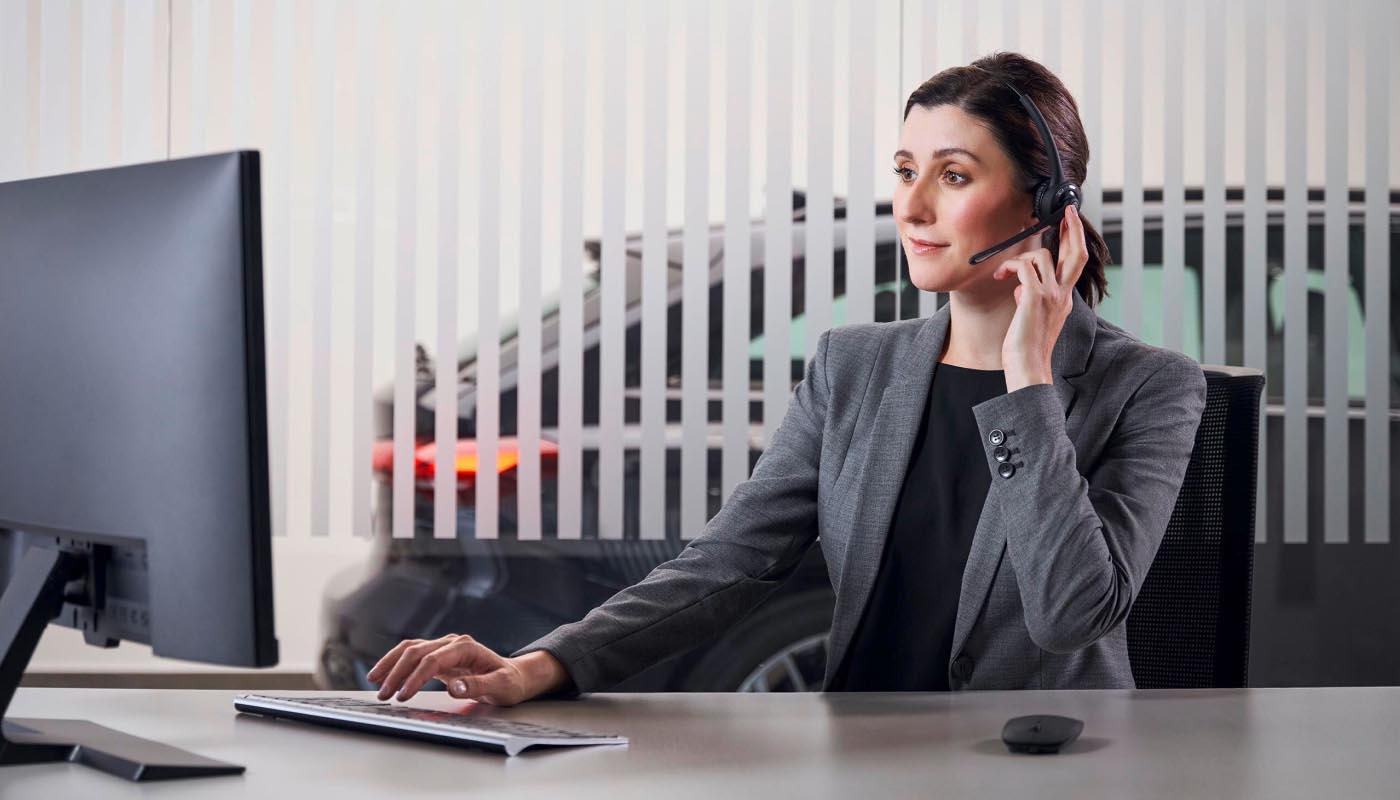 Eine Frau im Business-Outfit sitzt an ihrem Schreibtisch und telefoniert. Durch das Fenster des Büros ist ein Audi Q4 e-tron SUV zu erkennen.